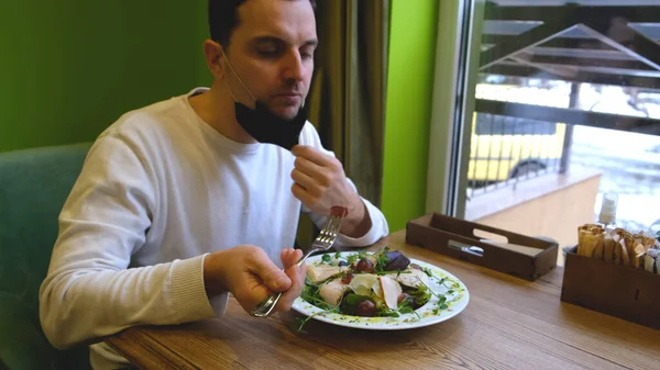 Un hombre se sienta en una mesa en un restaurante con una máscara, baja la máscara pone comida en su boca y pone la máscara de nuevo — Foto de Stock