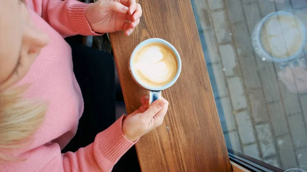 Uma menina loira senta-se em um café em frente à janela olha para a distância e bebe café de uma xícara de papel — Fotografia de Stock
