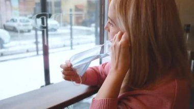 Sarışın, beyaz bir kız pencerenin karşısındaki kafede oturuyor ve maske takıyor.