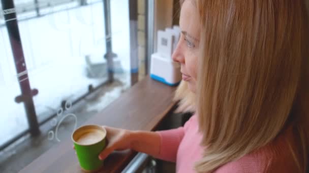 Блондинка сидить у кафе навпроти вікна дивиться на відстань і п'є каву з паперової чашки — стокове відео