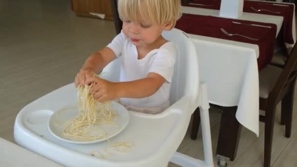 Baby boy eats spaghetti sitting in a baby chair in restaurant — 图库视频影像