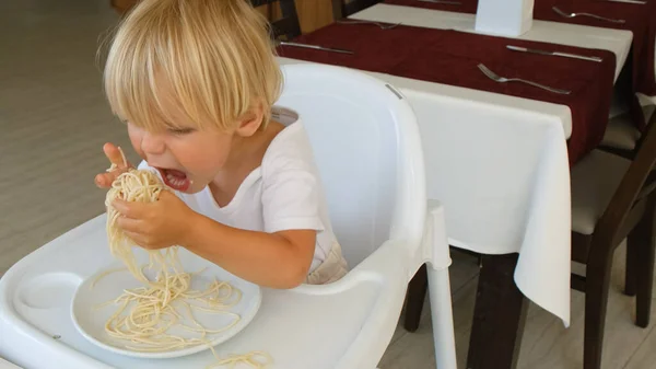 Baby boy eats spaghetti sitting in a baby chair in restaurant — Fotografia de Stock