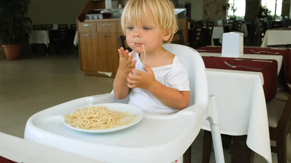 Baby boy eats spaghetti sitting in a baby chair in restaurant — Fotografia de Stock
