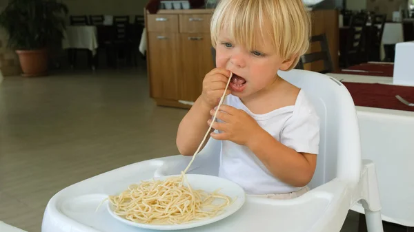 Baby boy eats spaghetti sitting in a baby chair in restaurant — Foto Stock