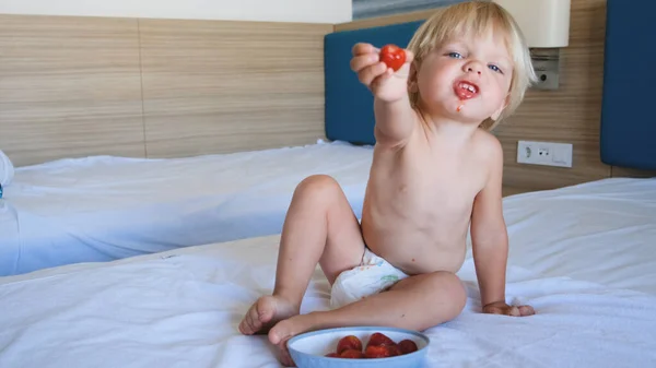 A little boy sits on a bed and pulls a hand with a strawberry forward — Fotografia de Stock