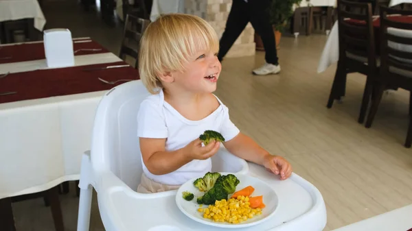 A little boy sits in a highchair and eats broccoli — Zdjęcie stockowe