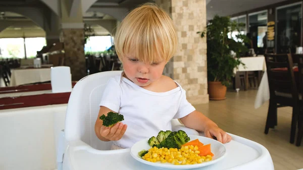 A little boy sits in a highchair and eats broccoli — Fotografia de Stock