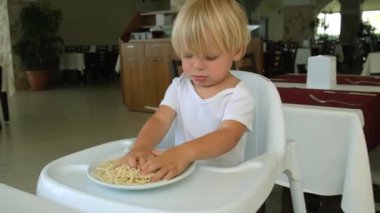 Baby boy eats spaghetti sitting in a baby chair in restaurant