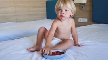 Küçük bir çocuk yatağa oturur ve çilek yer.