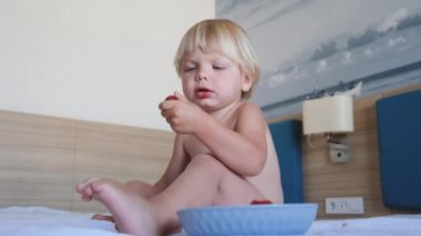 Küçük bir çocuk yatağa oturur ve çilek yer.