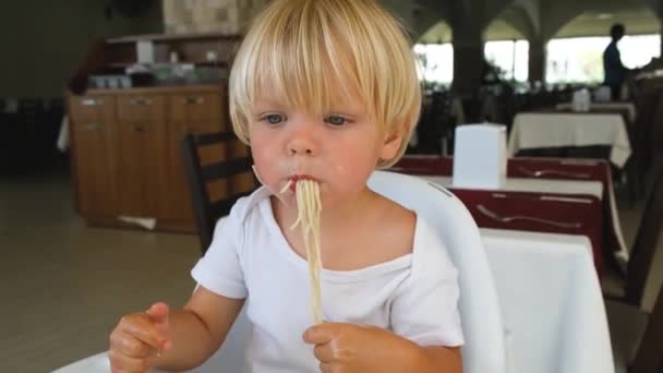 Baby boy eats spaghetti sitting in a baby chair in restaurant — 图库视频影像