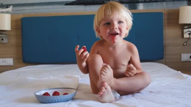A little boy sits on a bed and eats strawberries — 图库视频影像