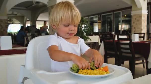 Маленький мальчик сидит в кресле и ест овощи — стоковое видео