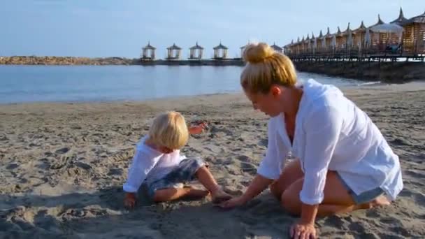 Pequeño niño en una camisa blanca juega en la arena en la playa con mamá — Vídeos de Stock