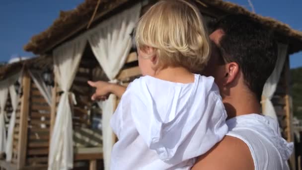 Padre sostiene a su hijo en sus brazos y le muestra algo con un dedo en camisas blancas — Vídeo de stock