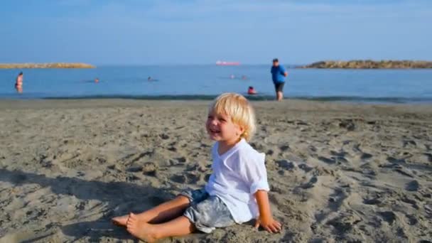 Menino em uma camisa branca joga na praia jogando areia na câmera — Vídeo de Stock