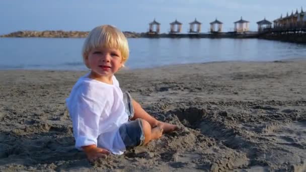Menino com uma camisa branca joga na areia na praia — Vídeo de Stock