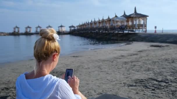 Uma menina em uma camisa branca leva uma selfie na praia — Vídeo de Stock