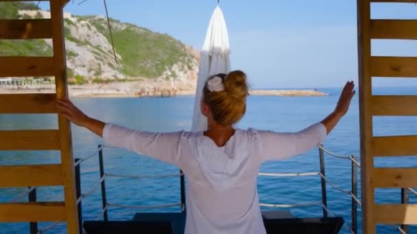 A girl in a white light shirt and with a white flower in her hair looks at the sea — Stock Video