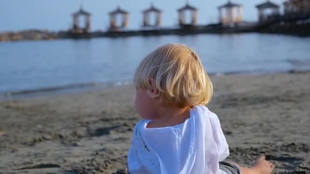 Menino em uma camisa branca senta-se na areia e olha para o mar — Vídeo de Stock