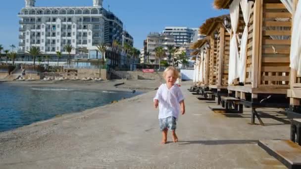 Мальчик в белой рубашке на берегу моря бежит к камере — стоковое видео