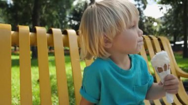 Parktaki sarı bankta oturan küçük bir çocuk dondurma külahı yiyor. Yüksek kalite 4k görüntü