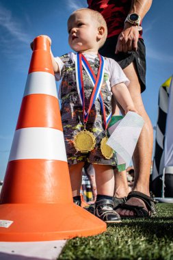 Küçük çocuk trafik konisinin yanında duruyor, onu elinde tutuyor. Boynumda iki altın madalya asılı.