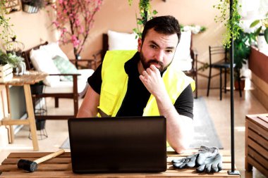 Çalışma alanında bir bilgisayar, eldiven ve çekiçle çalışan bir işçi.