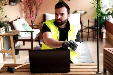 Çalışma alanında bir bilgisayar, eldiven ve çekiçle çalışan bir işçi.