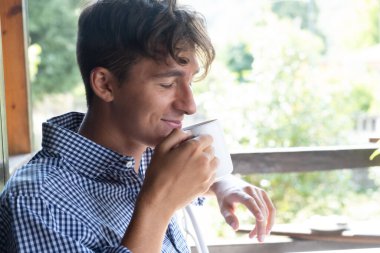 Genç bir adam doğada bir balkonda gülümseyip çay kokluyor. Sakin ve sakin bir atmosfer. Ergenlik. 