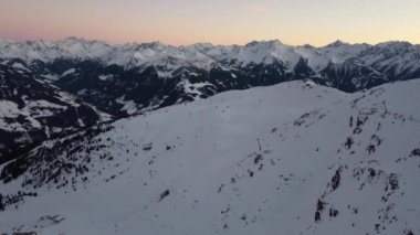 Snowy mountain ski-lifts and furniculars and ski-slopes in Austrian Alps drone flyover the mountains during sunrise golden hour orange sky