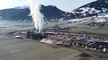 Wood sawmill proccessing wood logs and manufacturing wooden cuts and chimneys smoke is polluting the environment in beautiful natural set of deep valley surrounded by mountains