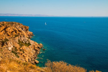 Akdeniz 'deki Cape Sounion' dan (Yunanistan) arka planda yelkenli ve ön planda turuncu kum kayalarıyla doğuya bakan görüntüler