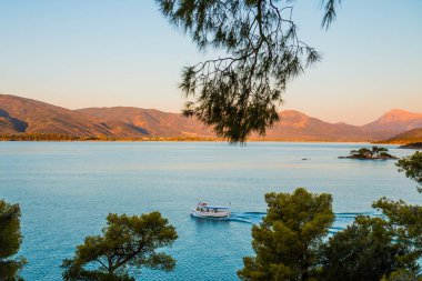 Arka planda küçük bir ada ve yarım ada olan Yunanistan 'ın Poros körfezinden kalkan küçük bir teknenin görüntüsü ve güzel mavi su rengi ve günbatımı ışıkları.