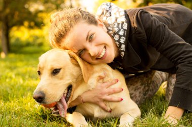 Gülümseyen bir kız parkta köpeğiyle oynuyor.