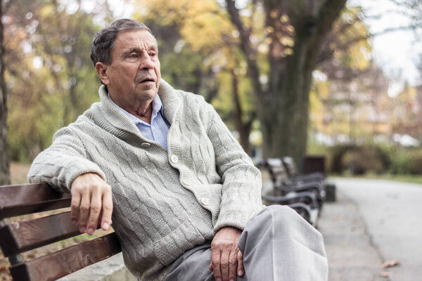 Portrait of a pensive senior man sitting on the bench, in the public park, outdoors. Old man relaxing outdoors and looking away. Portrait of elderly man enjoying retirement