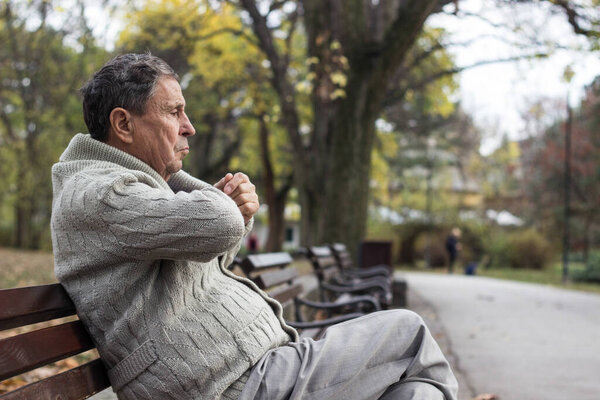 Portrait of a pensive senior man sitting on the bench, in the public park, outdoors. Old man relaxing outdoors and looking away. Portrait of elderly man enjoying retirement