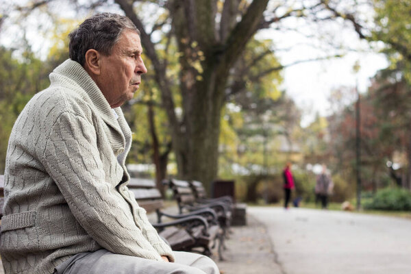 Portrait of a pensive senior man sitting on the bench, in the public park, outdoors. Old man relaxing outdoors and looking away. Portrait of elderly man enjoying retirement