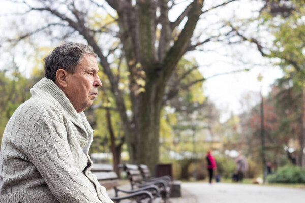 Portrait of a pensive senior man sitting on the bench, in the public park, outdoors. Old man relaxing outdoors and looking away. Portrait of elderly man enjoying retirement