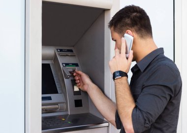 Genç adam telefonda konuşuyor ve kredi kartını bankamatiğe koyuyor.
