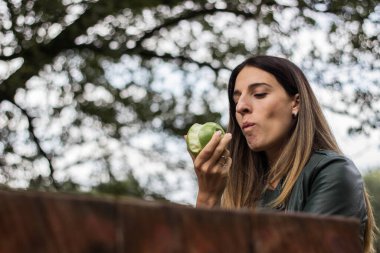 Halka açık parkta elma yiyen neşeli genç bir kadın. Oturmak, gülümsemek ve eğlenmek.