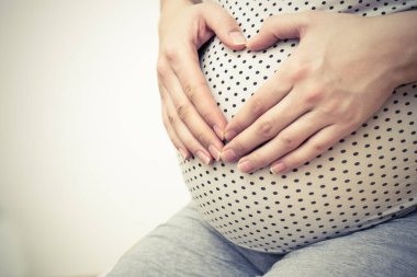 Happy pregnant woman making heart gesture. Holds hands on belly at home interiors.