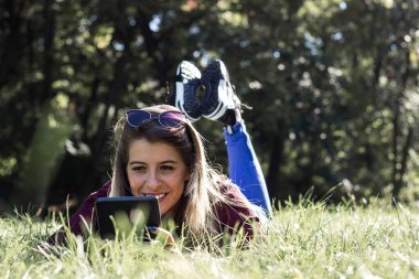 Gülümseyen kadın çimlere uzanıyor ve tablet kullanıyor.