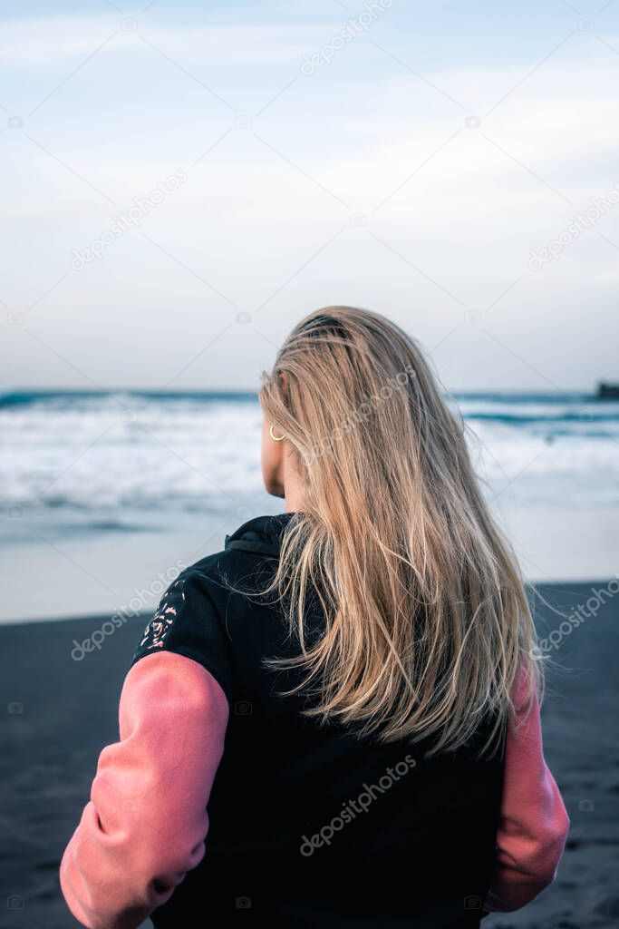 Unrecognizable blonde girl seen from behind looking at the sea on a black sand beach