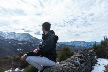 Arkadan görülen genç adam doğada oturmuş manzarayı seyrediyor.