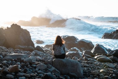 Arkadan kumsalda oturmuş günbatımını izlerken görülen kahverengi saçlı kadın. Arka planda dalgalar çarpıyor.