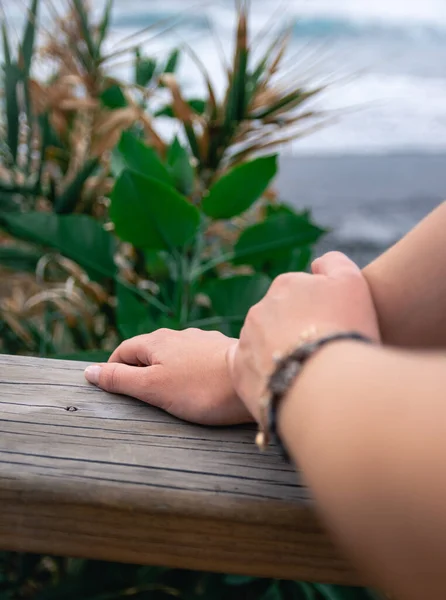 Крупный план белокожих женских рук, лежащих на деревянных перилах перед морем и немного растительности — стоковое фото