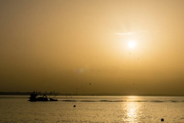 Bright sunrise with large yellow sun with rays under the sea surface wit silhouette of birds and boat, Lagos, Portugal