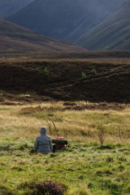 Çayırda oturan bir kadın yürüyüş sonrası İskoç manzarasına bakıyor.
