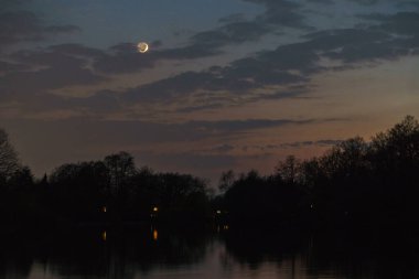 Günbatımından sonra hilal renkli bir gökyüzünde göl kıyısındaki ağaçların üzerinde
